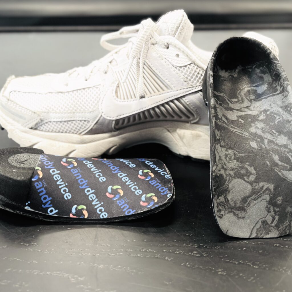 “Custom pediatric orthotics with black marbled support surface and branded top layer displayed alongside a white athletic shoe.”